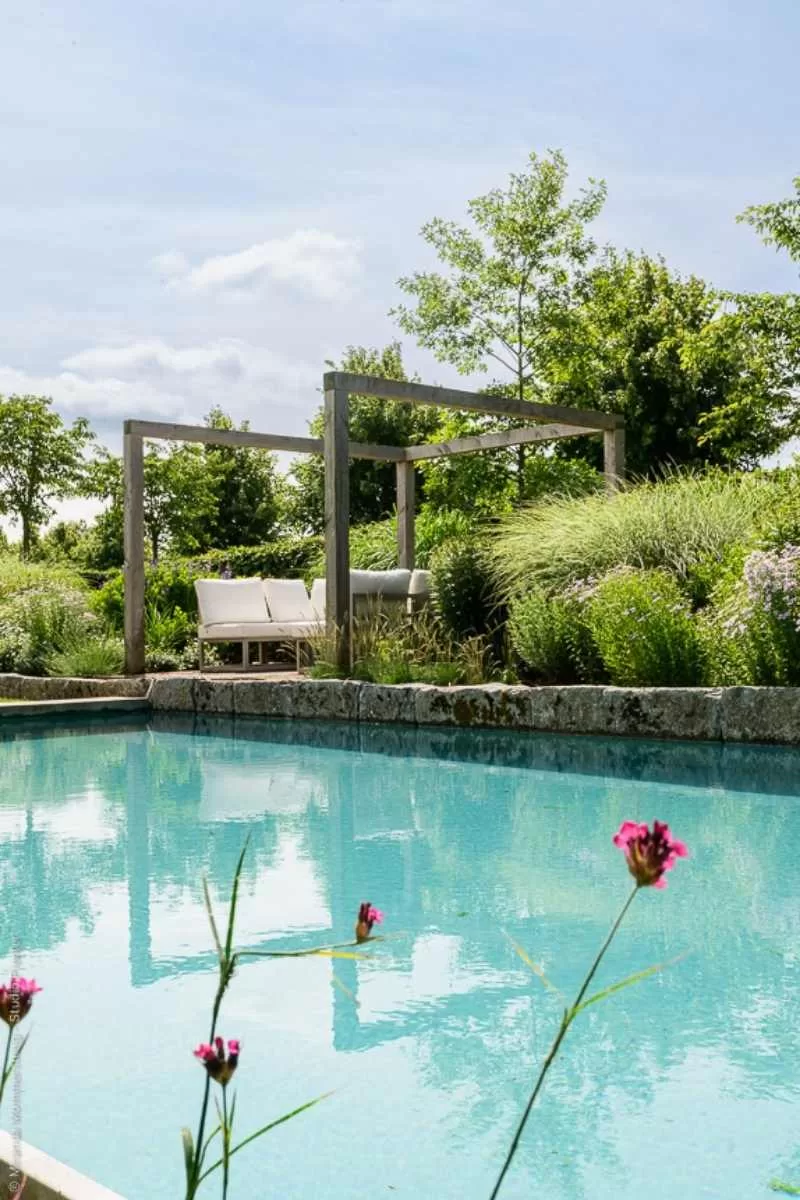 Helderblauwe zwemvijver met robuuste natuurstenen rand en houten pergola in groene tuin